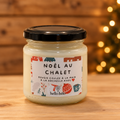 Bougie artisanale Noël au chalet, pot en verre fermé avec étiquette illustrée de motifs de Noël, sur une table en bois devant des lumières dorées