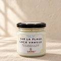 Bougie artisanale Sur la plage coco vanille, pot en verre fermé avec étiquette illustrée, sur un lin beige devant un mur crème éclairé par la fenêtre